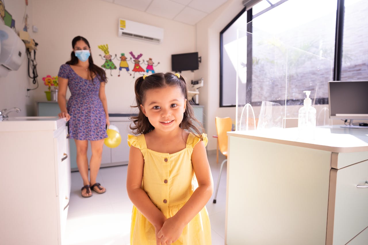 about-04 A cheerful child in a yellow dress at a pediatric clinic with doctor in background.